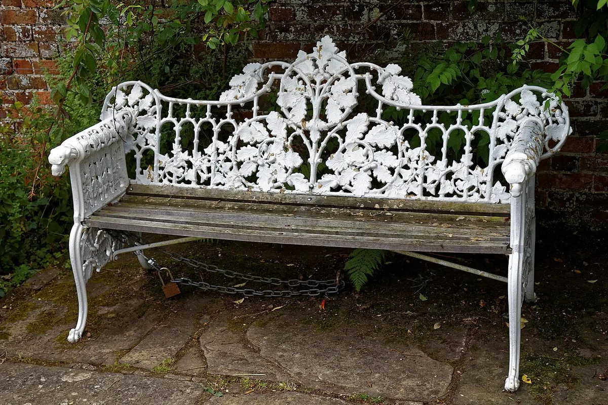 Verwittertes ornamentales Gusseisenbänkchen mit Blattmotiven vor einer Backsteinmauer im Garten